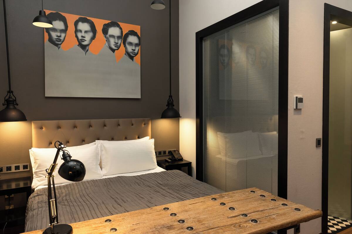 A modern hotel room with a tufted headboard, a wooden bedside table, a large framed artwork above the bed, and a sliding glass door to the right.