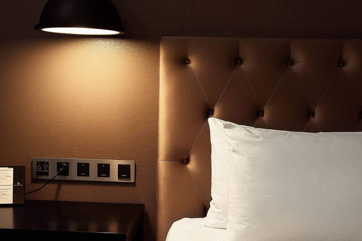 A neatly made bed with a white pillow and a striped blanket, a dark wooden nightstand with a lamp, and a wall-mounted power outlet.
