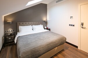 Quarto moderno com cama, mesas de cabeceira, termostato fixado na parede e uma porta.