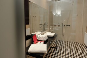 Uma casa de banho moderna com chuveiro de vidro, chão em padrão xadrez preto e branco e toucador com uma caixa de produtos de higiene pessoal vermelha.