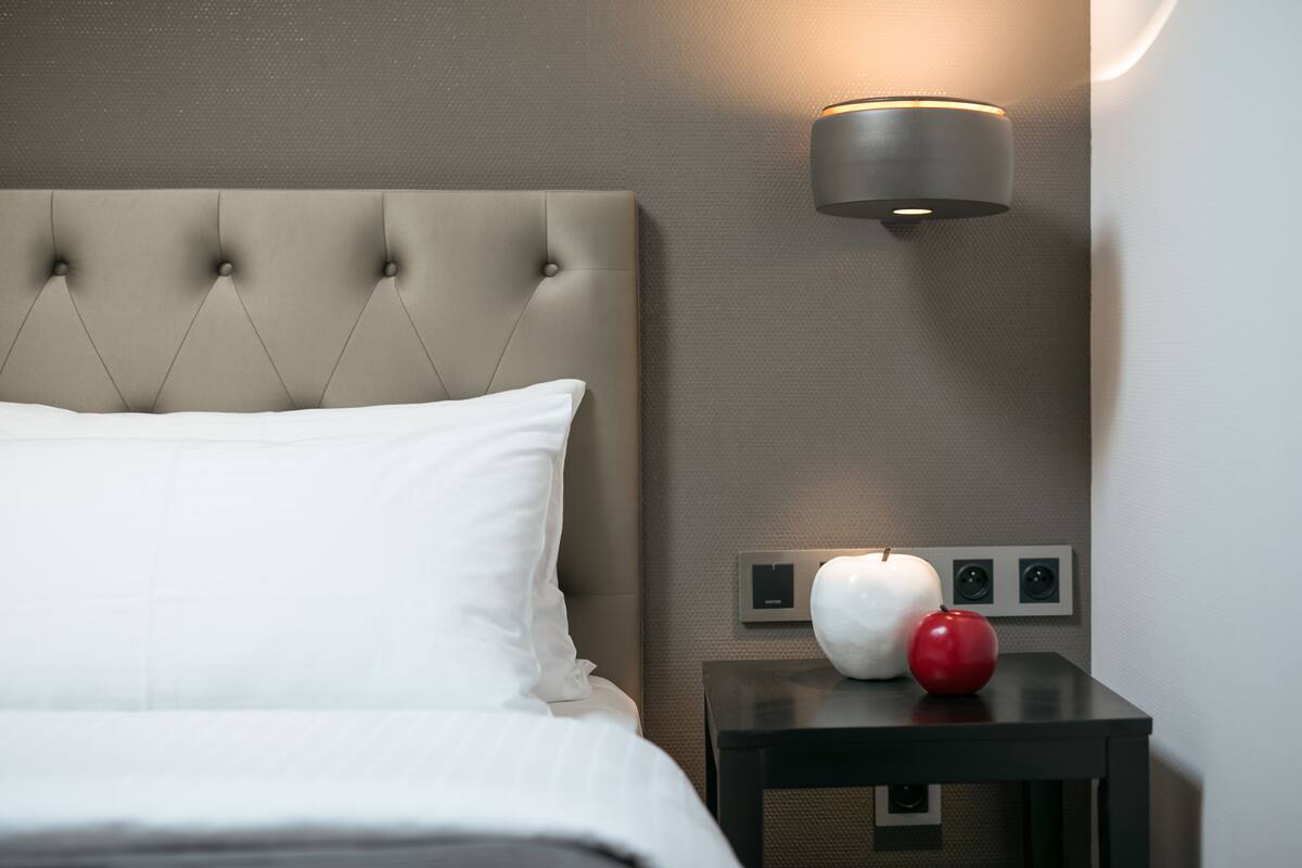 A neatly made bed with a tufted headboard, a bedside table with a white apple and a red apple, and a wall-mounted light fixture.