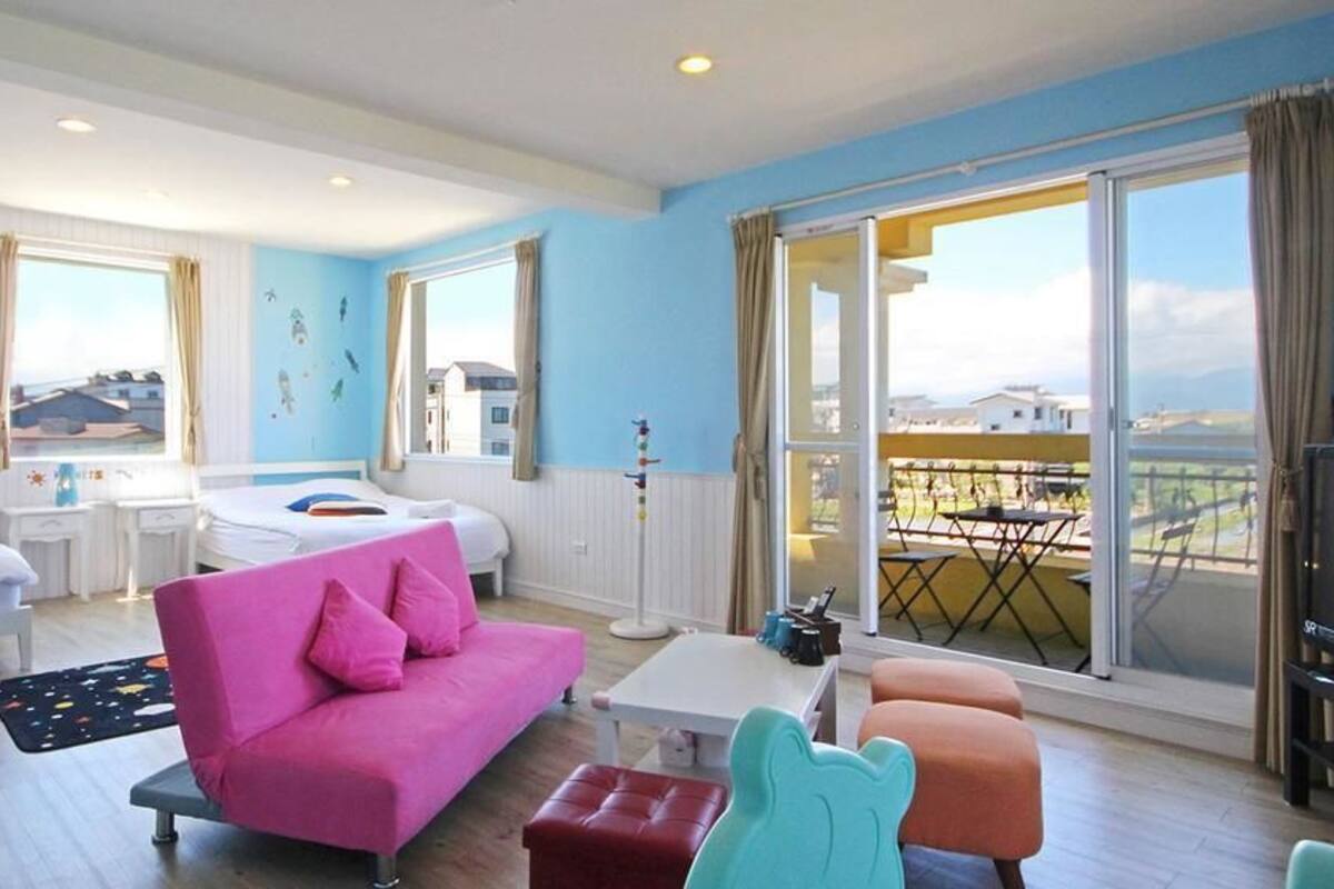 A bright room with a pink sofa, two beds, a table, and a balcony with a view.