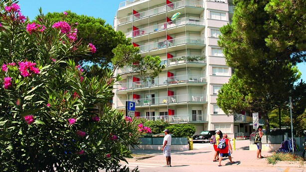 A multi-story hotel with balconies, a parking sign, and lush greenery. - La Zattera (Lignano Sabbiadoro)