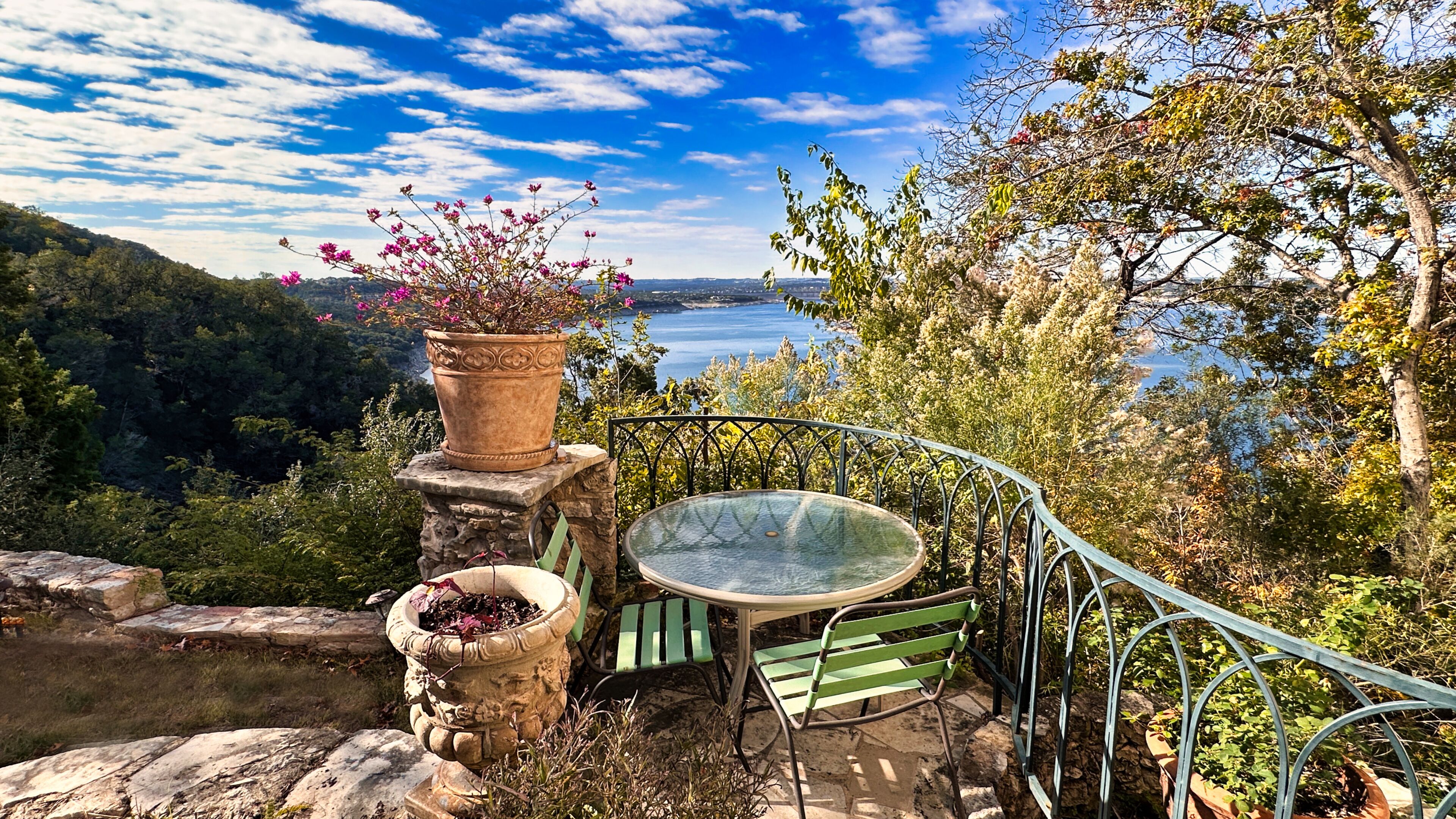 A patio with a table and chairs, surrounded by stone structures and potted plants, overlooking a scenic landscape with trees and a body of water.