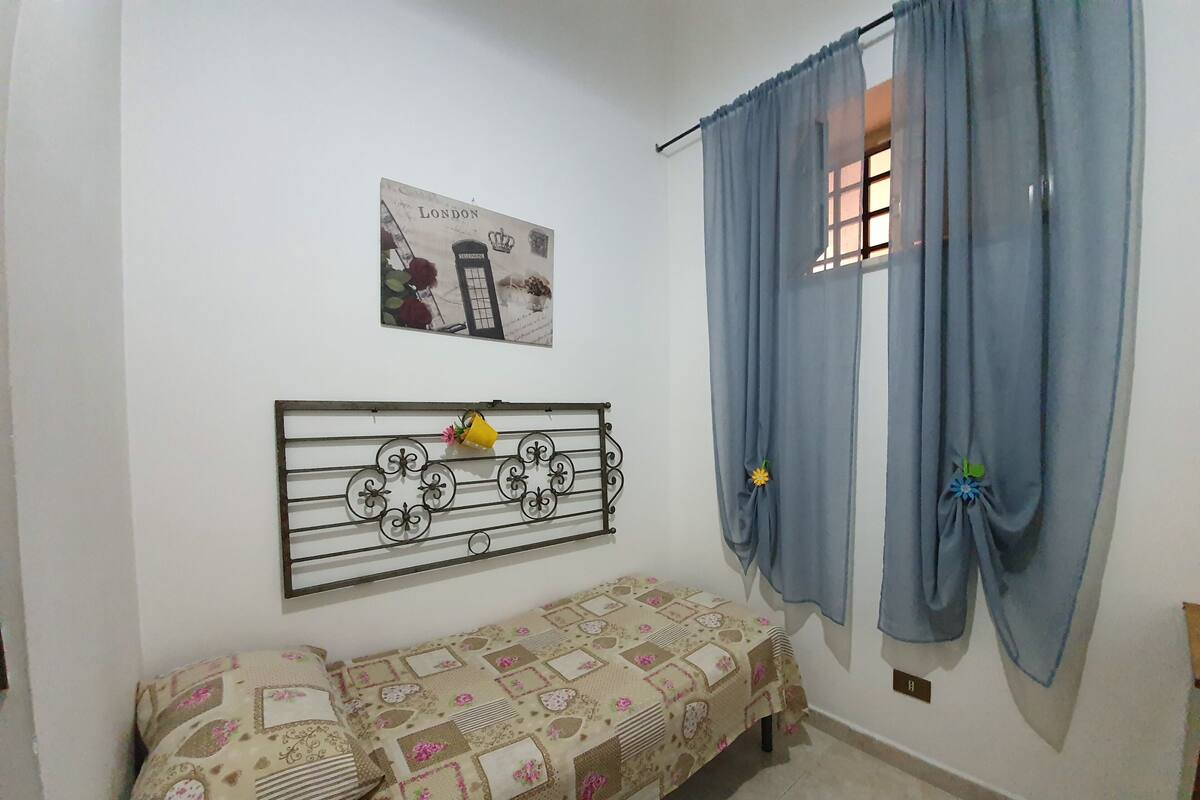 A small, cozy room with a single bed, a wall-mounted shelf, and a window with curtains.