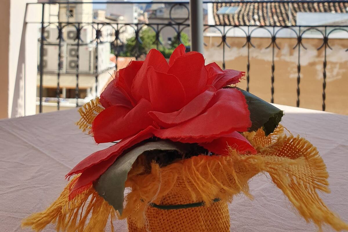 A balcony with a table, a decorative item with a red flower, and a view of buildings and mountains in the background.
