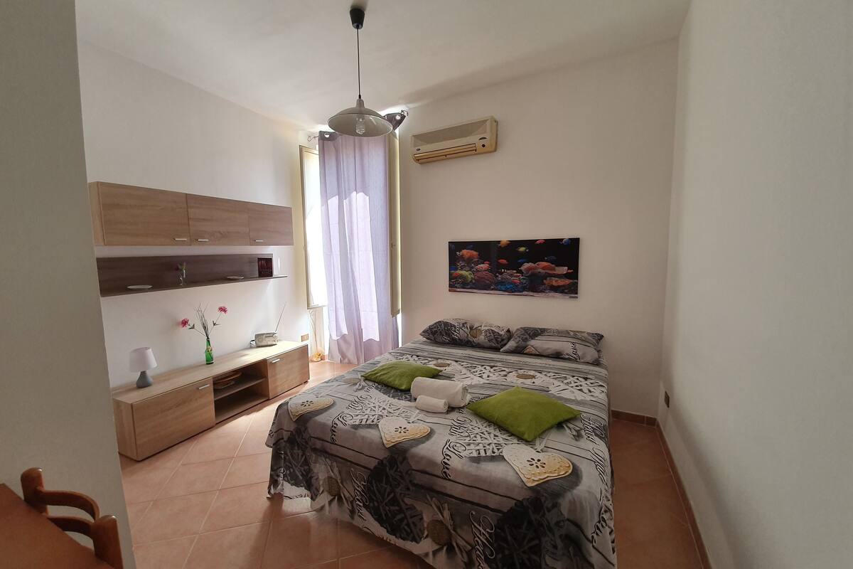 A bedroom with a bed, a nightstand, a lamp, a radio, a picture on the wall, and a window with curtains.