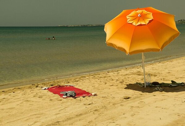 En strand med ett stort orangefärgat parasoll, en röd handduk och ett par skor som ligger på sanden.