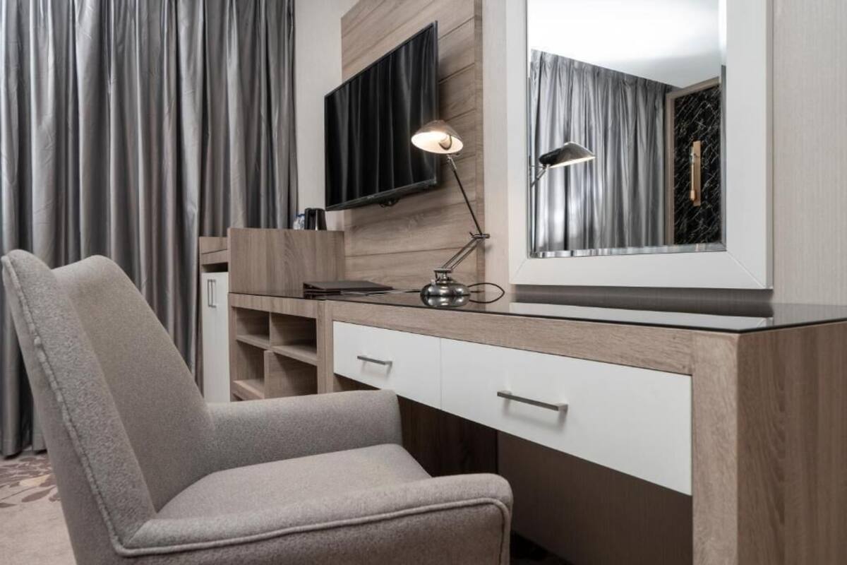 A modern bedroom with a wooden desk, a grey chair, a large mirror, and a television mounted on the wall.