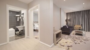 Moderni hotellihuone, jossa on kylpyhuone, oleskelutila sohvineen ja nojatuolineen sekä suuri ikkuna, jossa on verhot.