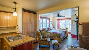 A spacious room with a large bed, a sitting area with a chair and ottoman, a kitchen with wooden cabinets, and a bathroom with a bathtub.
