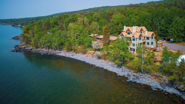 A large, multi-story house with a prominent roof, situated on a rocky shoreline with a forested hill in the background.