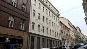 File di auto parcheggiate lungo una strada con un edificio dotato di numerose finestre e un cartello per 'Barberland'.