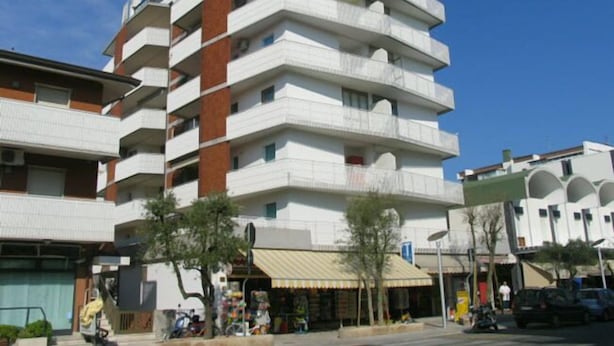 A multi-story building with a white facade and red accents, featuring balconies and a ground-level storefront. - Mediterraneo (Lignano Sabbiadoro)