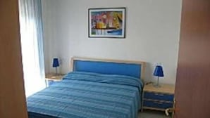 A bedroom with a bed, two bedside tables, a nightstand, a wall-mounted picture, and a window with curtains. - Mediterraneo (Lignano Sabbiadoro)