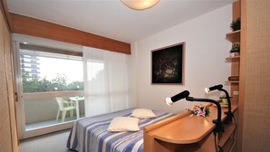 A bedroom with a bed, a desk with a lamp, a window with a view, and a painting on the wall. - Palme Residence (Lignano Sabbiadoro)