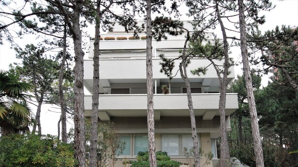 A modern multi-story building with a flat roof, surrounded by tall trees and dense shrubbery. - Palme Residence (Lignano Sabbiadoro)