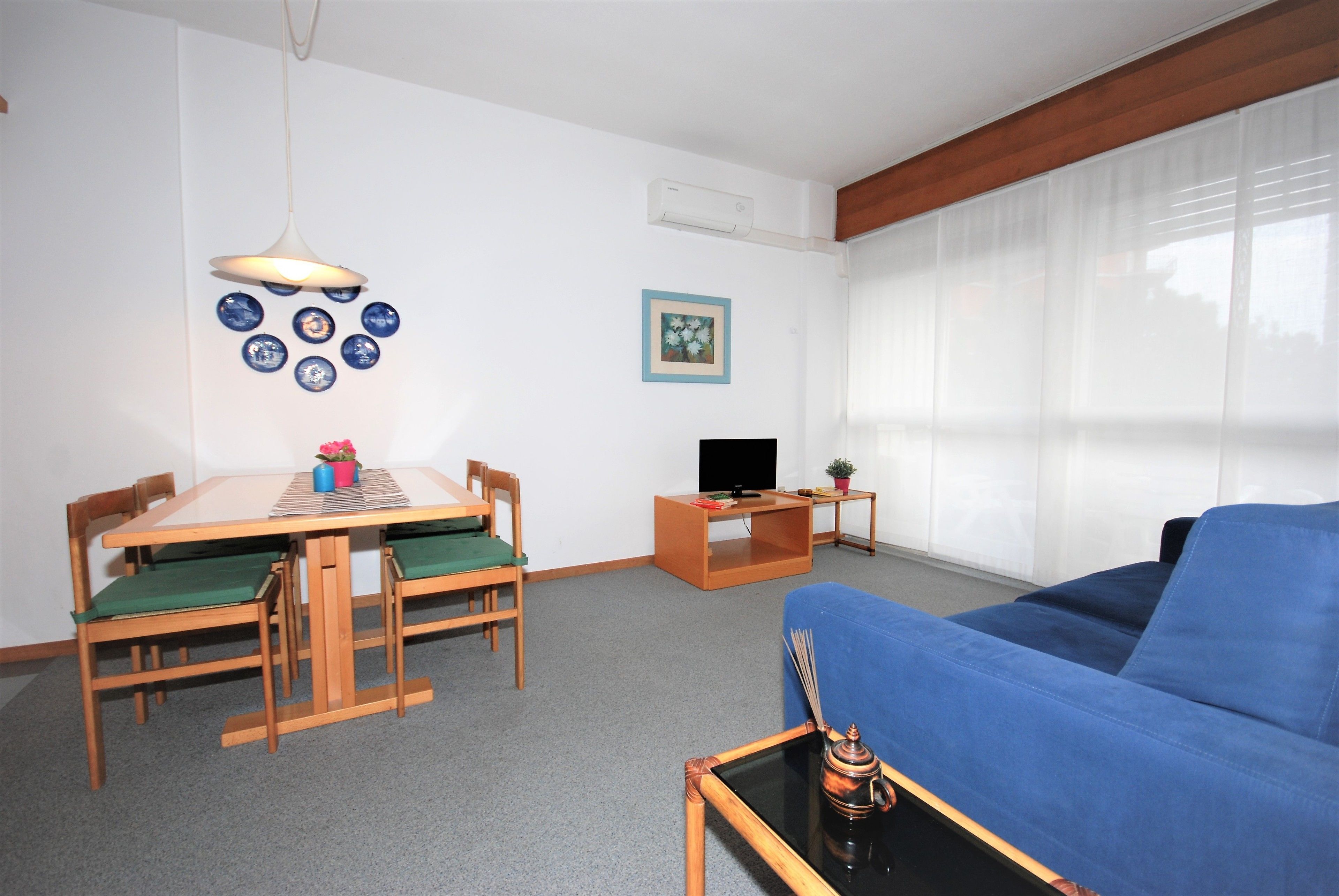 Een woonkamer met een blauwe bank, een houten eettafel met stoelen, een tv-meubel en een bijzettafeltje met een decoratief object.