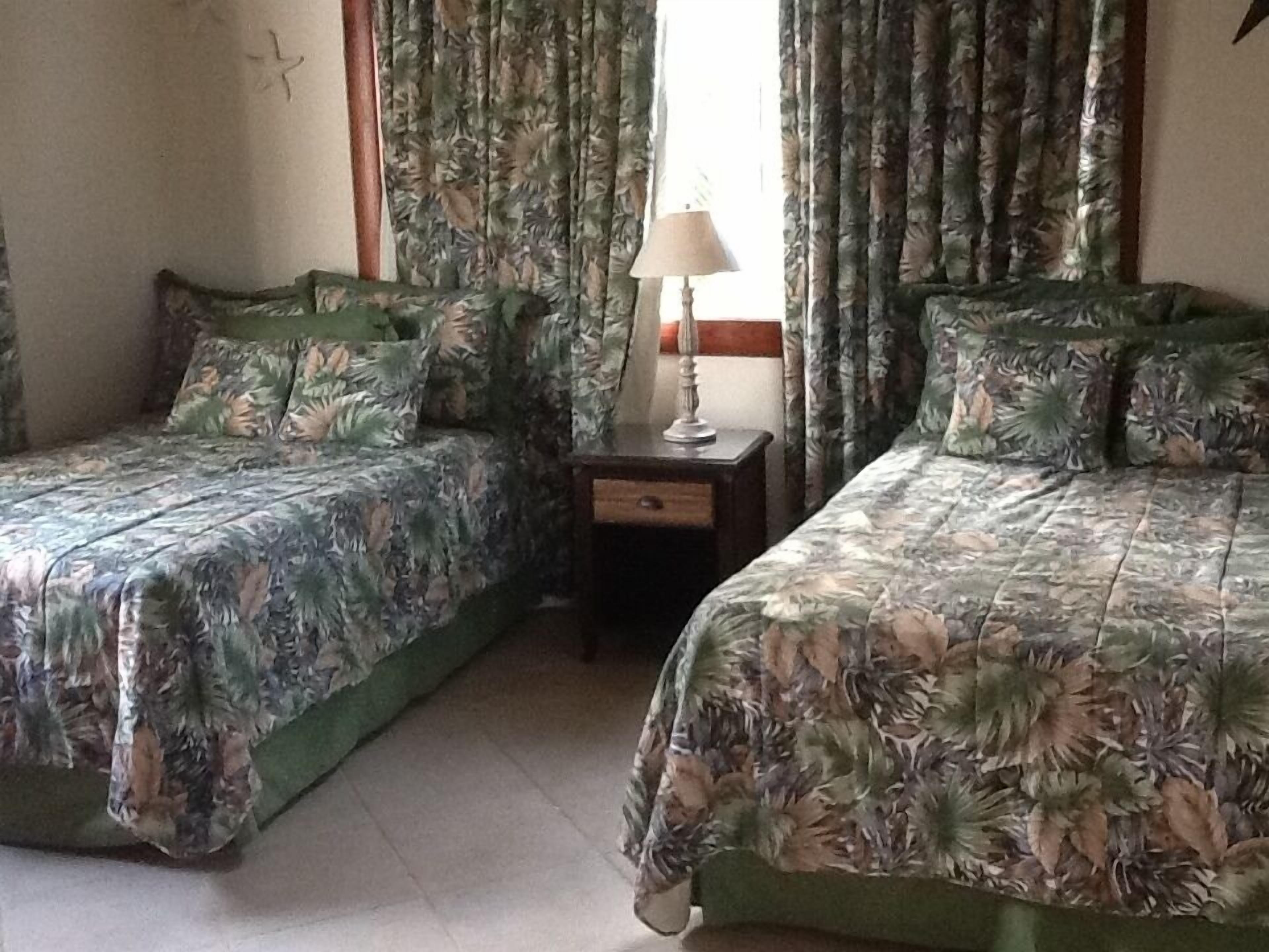 A room with two beds, each with floral-patterned bedding, a small bedside table with a lamp, and curtains with a leaf pattern.