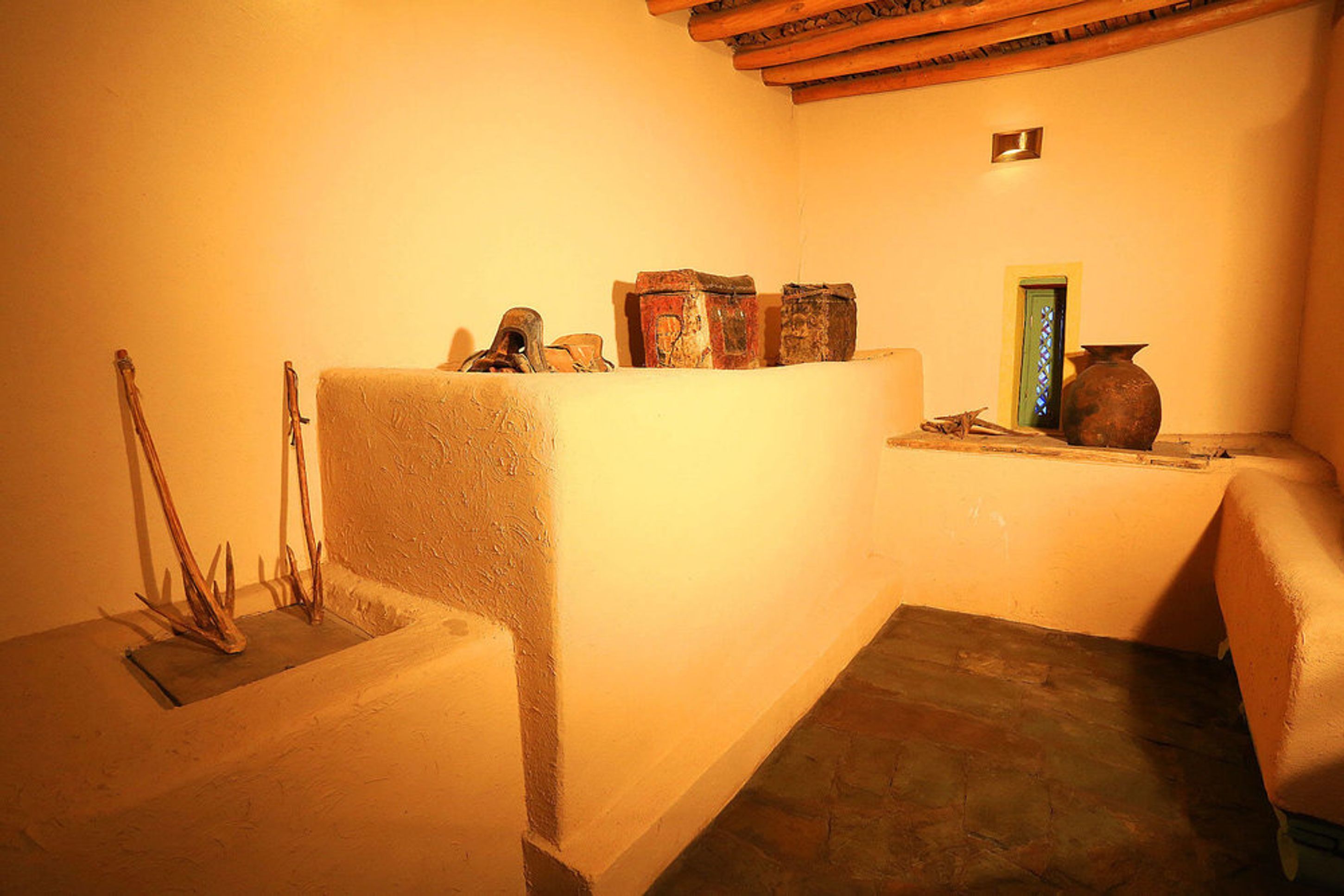 A corner of a room with a white wall, a wooden bench, and a small table with a vase and a door.