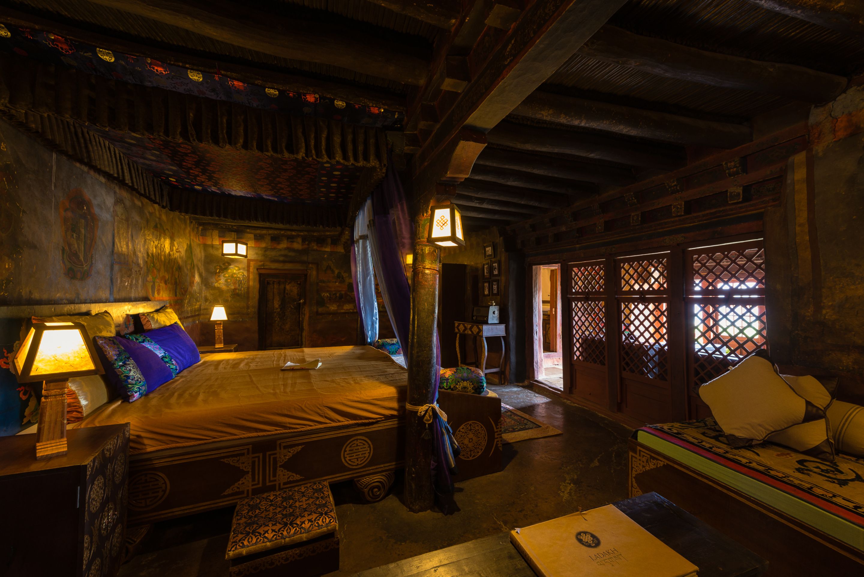 A traditional room with a bed, a sofa, and a table. There are lamps on the bedside table and a window with a lattice design.