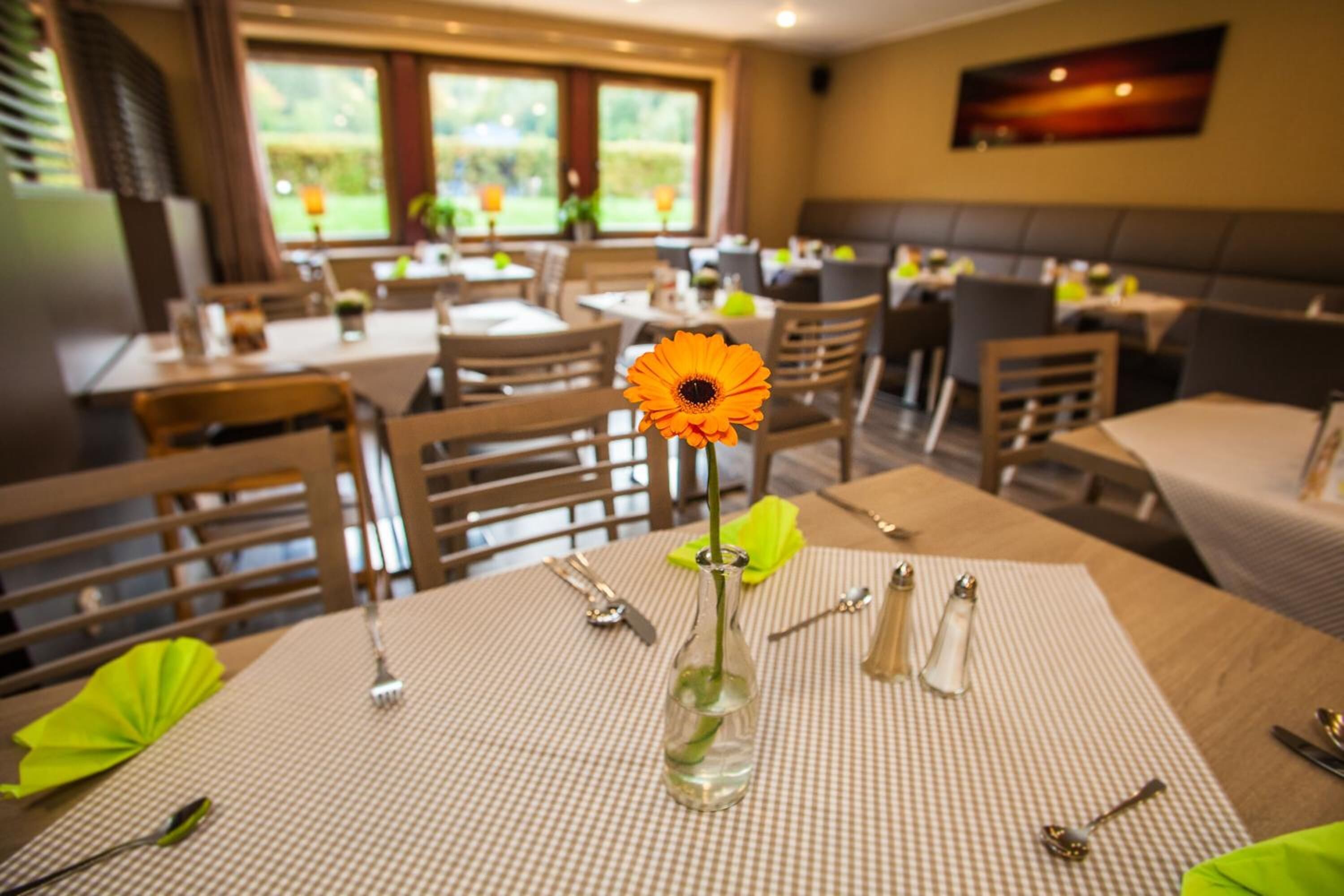 Un espace repas avec des tables dressées, un vase contenant une seule fleur et une vue sur l’extérieur grâce à de grandes fenêtres.