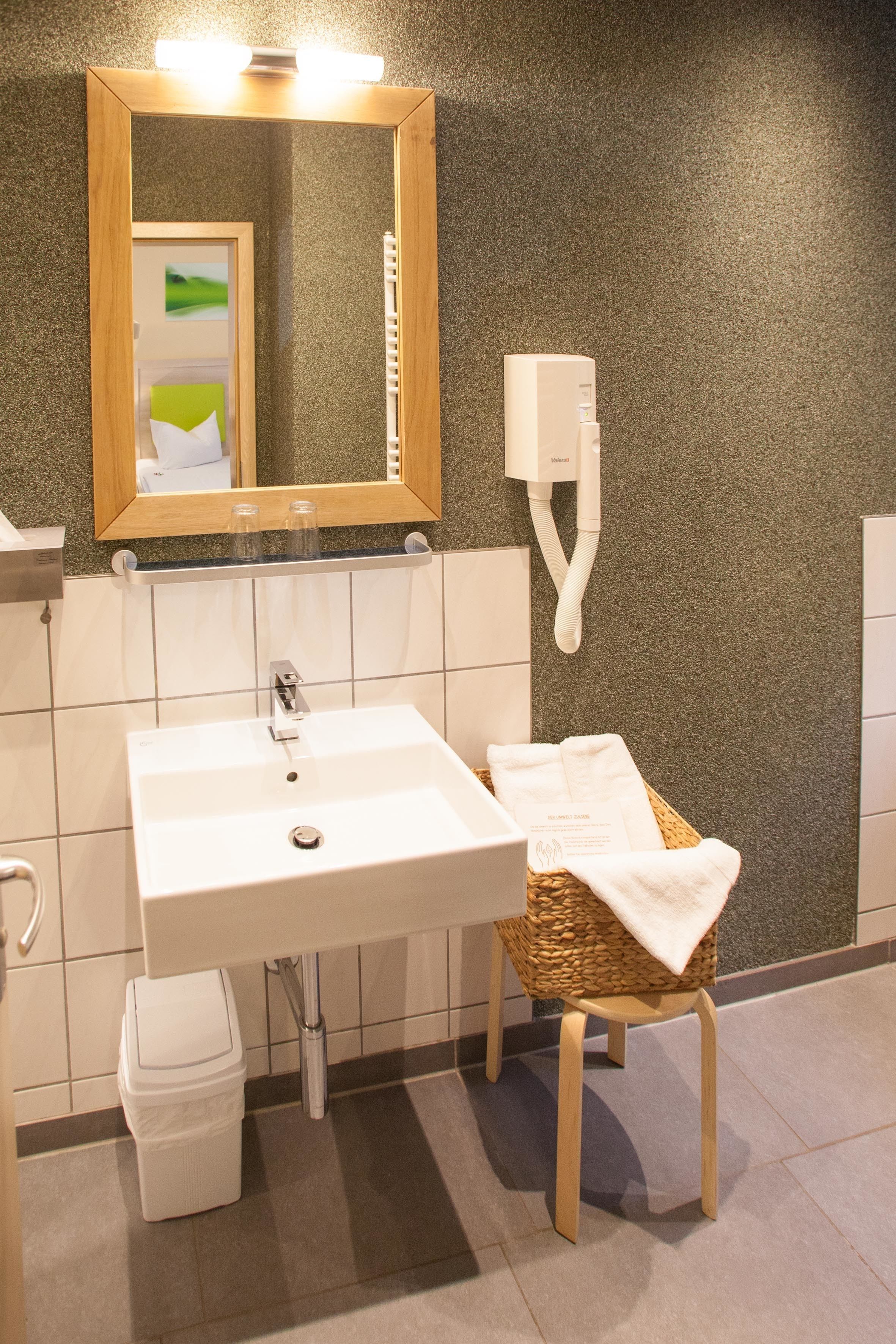 Une salle de bain avec un lavabo blanc, un miroir, un sèche-cheveux, une poubelle et une chaise en osier avec une serviette.