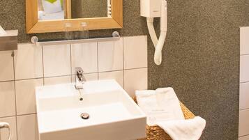 Une salle de bain avec un lavabo blanc, un miroir, un sèche-cheveux, une poubelle et une chaise en osier avec une serviette.