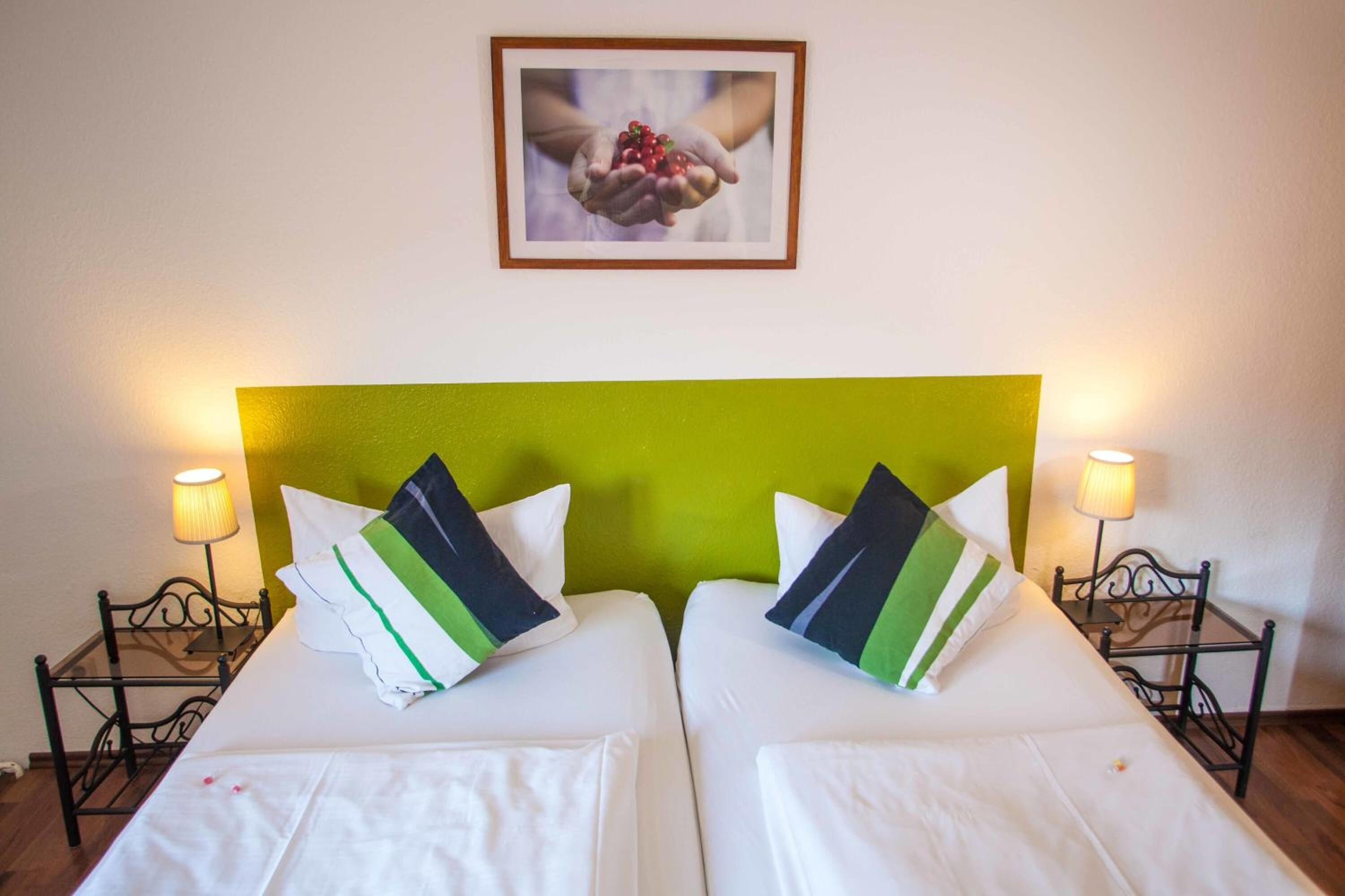 A hotel room with two beds, each with white linens and green and black striped pillows. There is a framed picture on the wall above the beds.