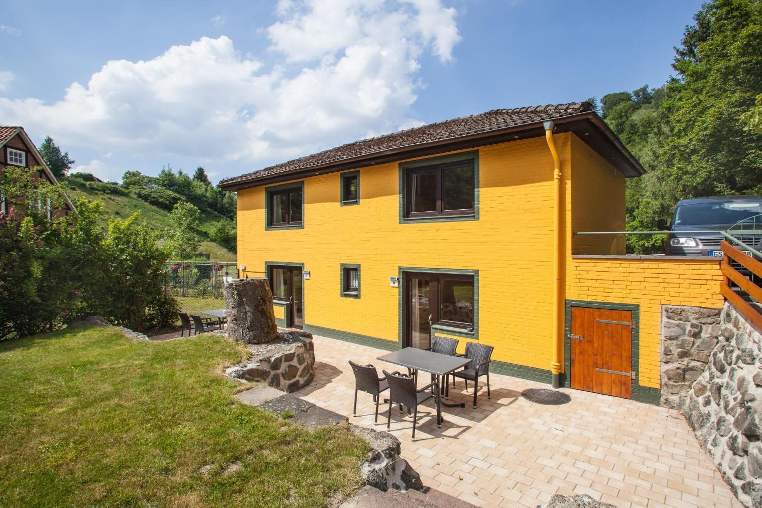 A two-story yellow building with a patio area featuring a table and chairs, a stone wall, and a car parked nearby.