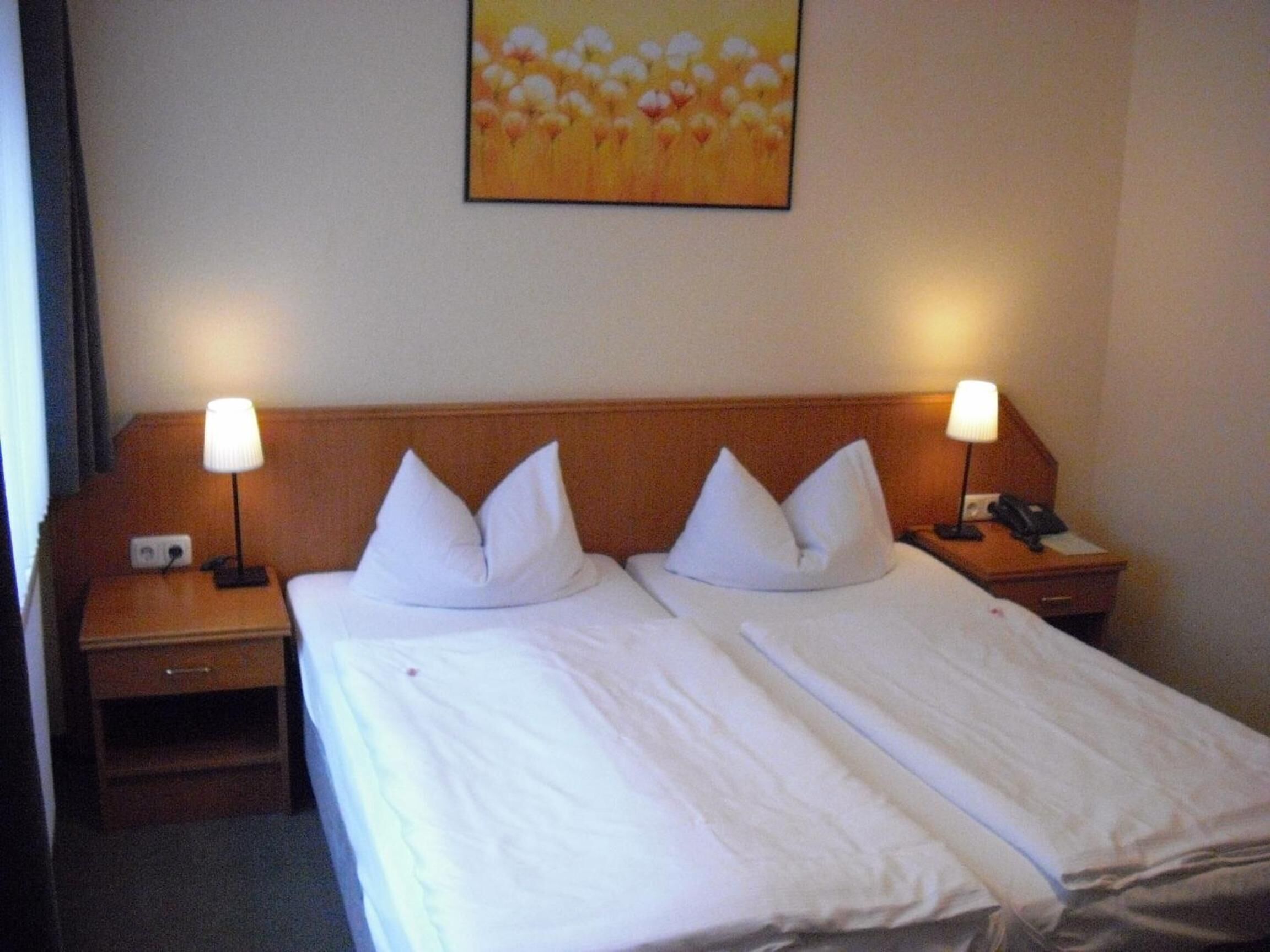 Habitación de hotel con dos camas, cada una con ropa blanca de cama y almohadas, flanqueadas por mesitas de noche de madera con lámparas y teléfono.