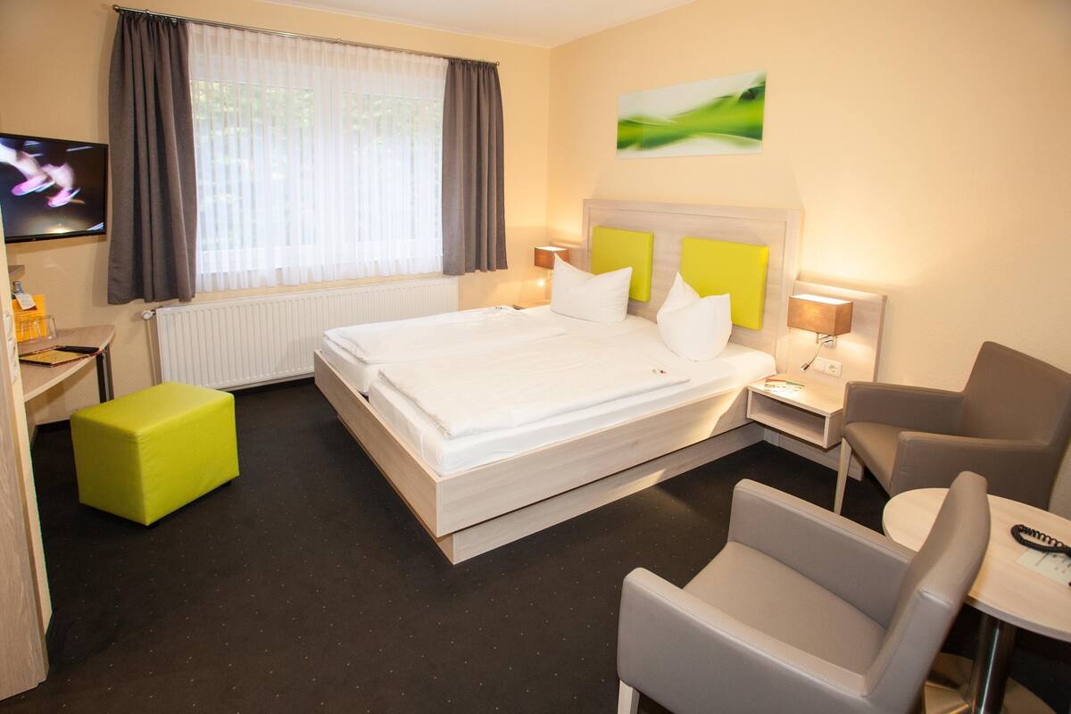 Ein Hotelzimmer mit einem Bett, einem grünen Hocker, einem Schreibtisch mit Stuhl und einem an der Wand montierten Fernseher.