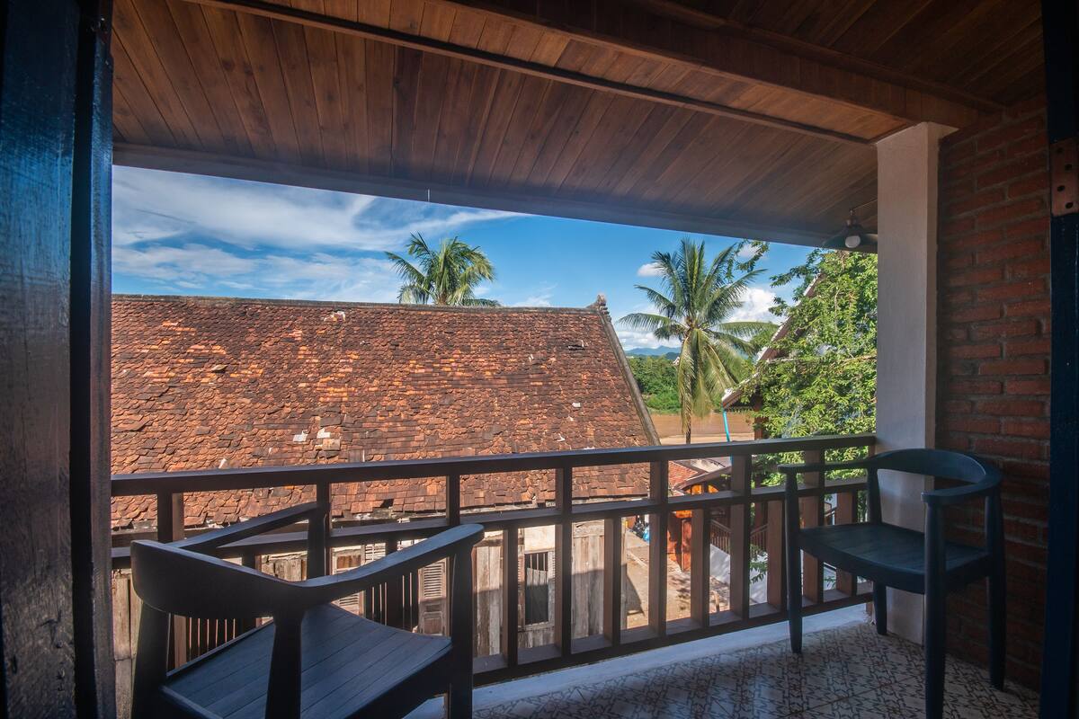 A balcony with wooden railings, a chair, and a view of a red-roofed building and palm trees.