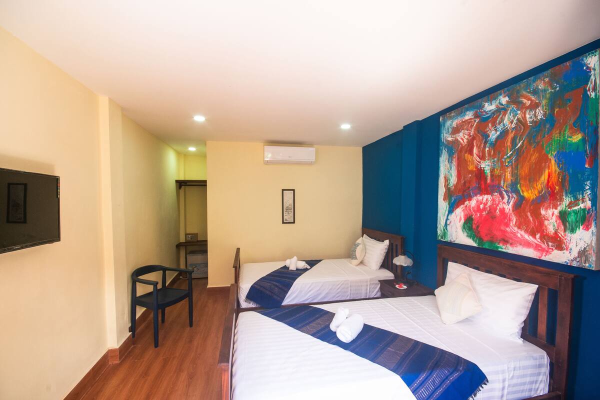 A hotel room with two beds, a chair, a TV, and a colorful abstract painting on the wall.