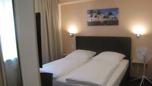 A hotel room with a bed, two bedside lamps, a nightstand, a fan, and a painting of palm trees on the wall. - Milano Hotel (Hamburg)