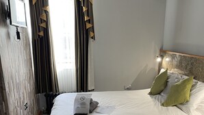 A hotel room with a large bed, a headboard with lights, a TV mounted on the wall, and a window with curtains. - Six Bells Hotels (Enfield)