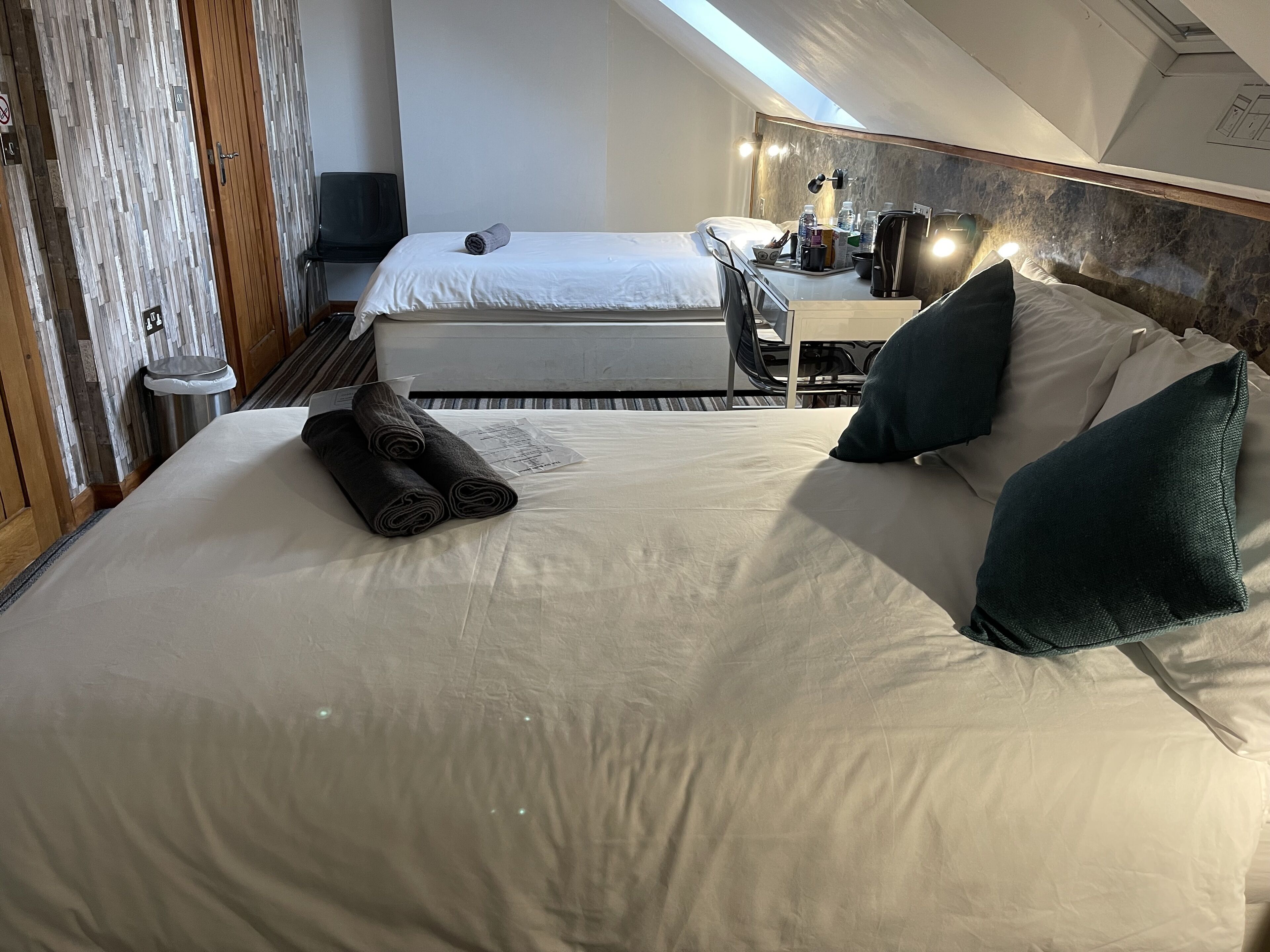 A hotel room with a large bed, a desk, and a chair. There is a towel on the bed and a bottle on the desk.
