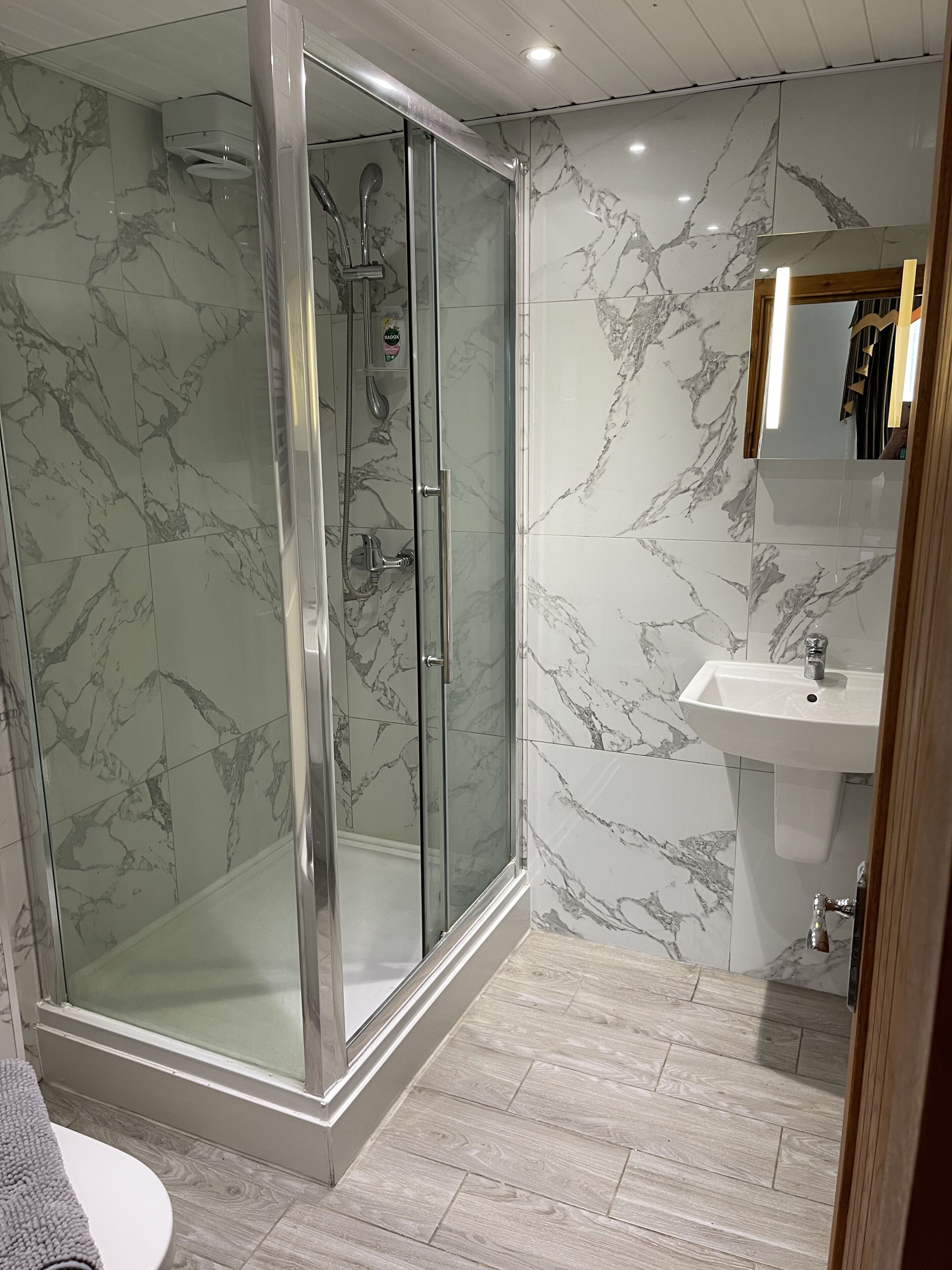 A modern bathroom with a glass-enclosed shower, marble-patterned walls, and a wooden floor.