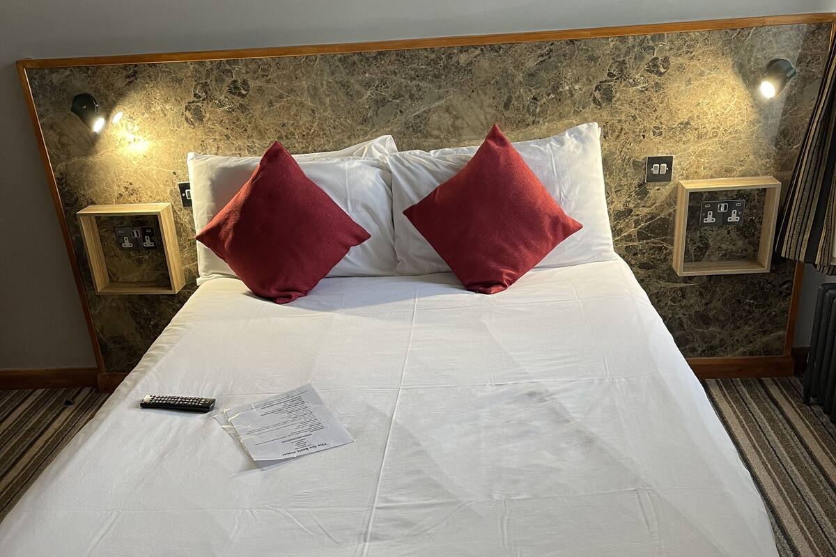 Ein ordentlich bezogenes Bett mit weißen Leinentüchern und zwei roten Kissen, einer Fernbedienung und einem Zettel darauf.