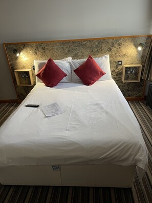Un lit bien fait, avec des draps blancs et deux oreillers rouges, une télécommande et un morceau de papier posés sur le lit.