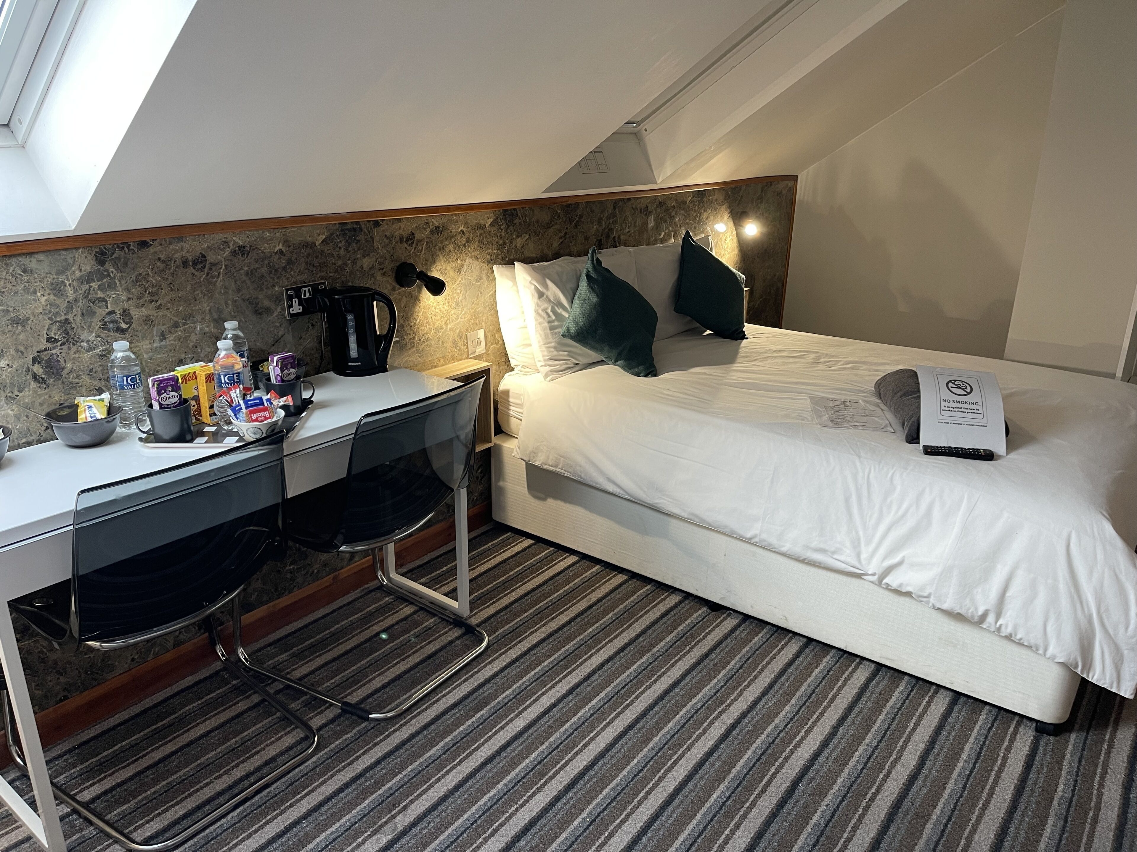 A hotel room with a bed, a desk, and a chair. There is a coffee maker on the desk and a sign on the bed.