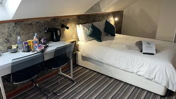 A hotel room with a bed, a desk, and a chair. There is a coffee maker on the desk and a sign on the bed.