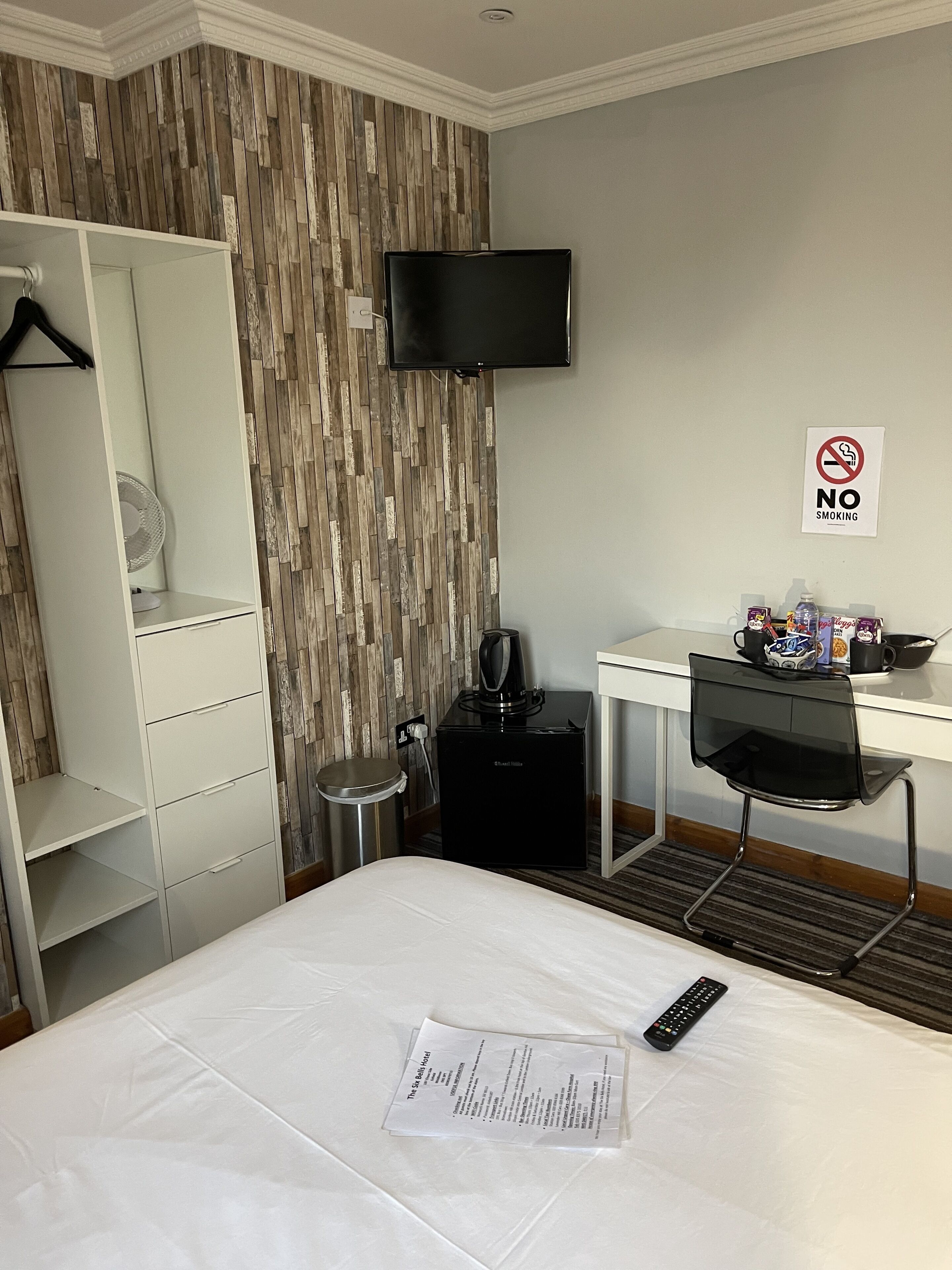 A hotel room with a bed, a desk, a chair, a TV, a coffee maker, a trash bin, and a no-smoking sign.