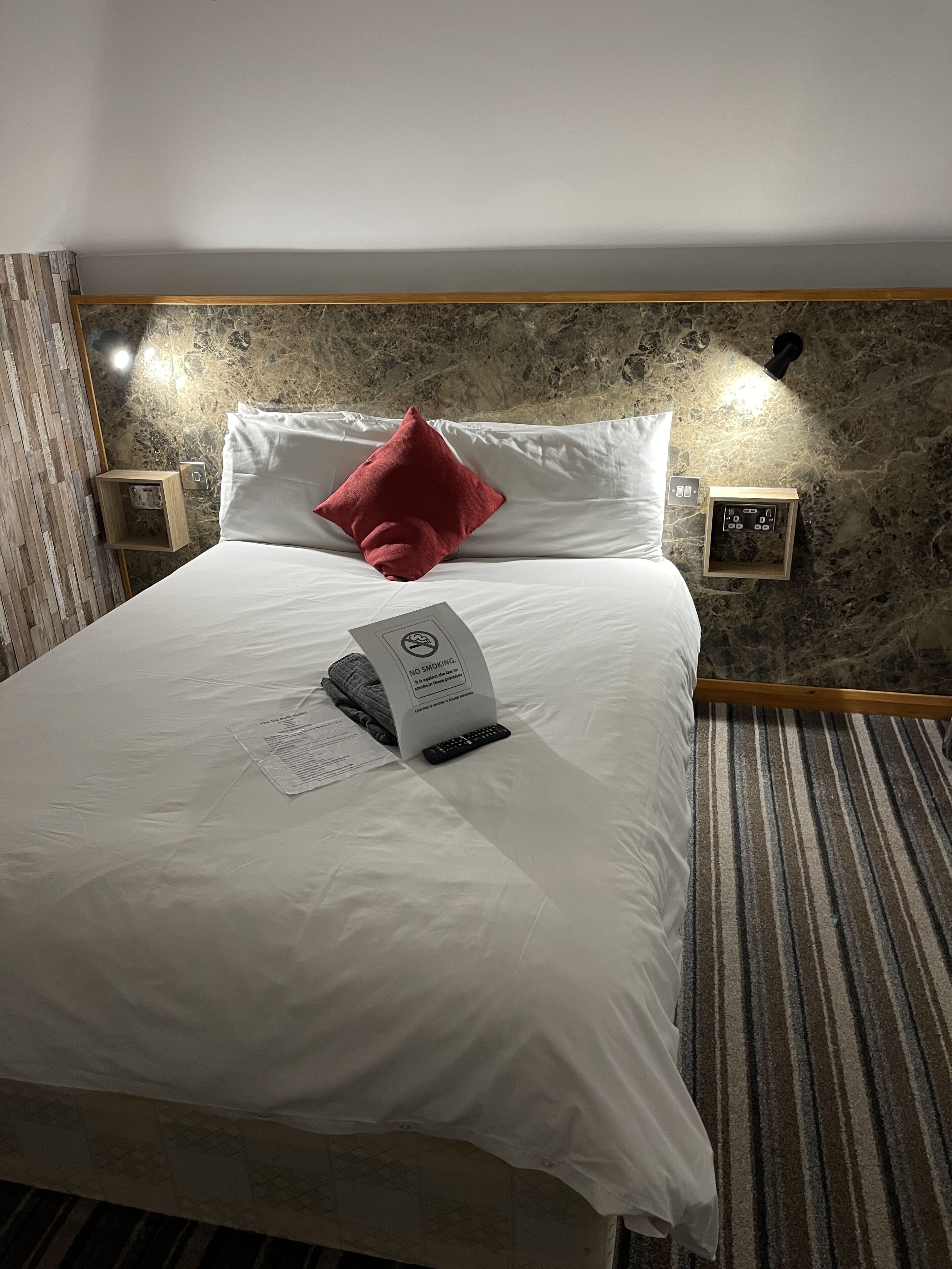 A neatly made bed with white linens and a red pillow, a remote control, and a no-smoking sign on the headboard.