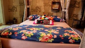Ein Bett mit blumigem Bettzeug und mehreren Kissen, ein Nachttisch und ein an der Wand befestigtes Regal.