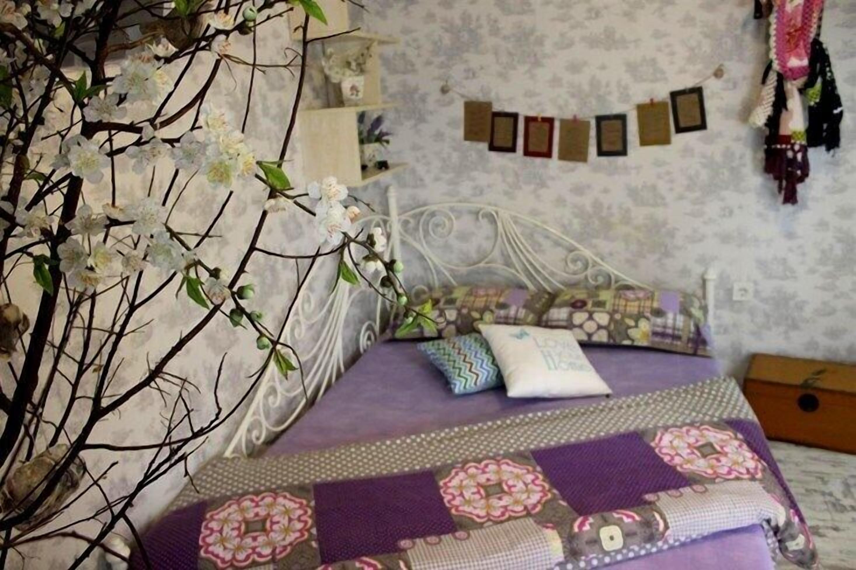 A bedroom with a bed, decorative pillows, a quilt, a wall with framed pictures, and a shelf with items.
