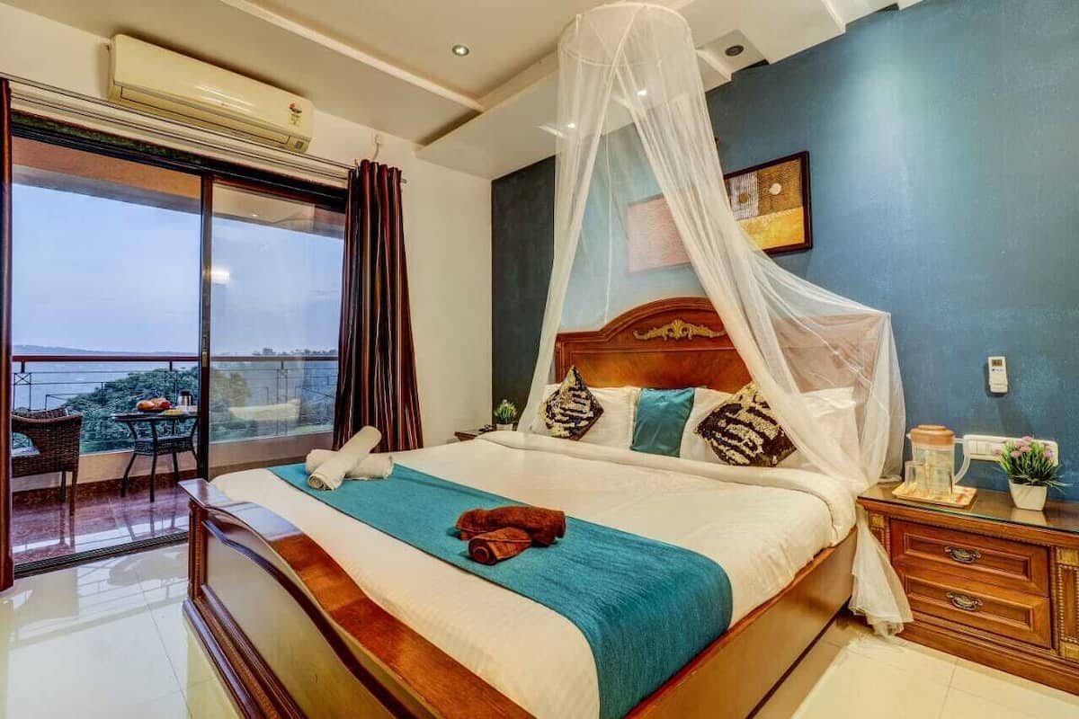 A bedroom with a large bed, a nightstand, a bedside table, a window with curtains, and a view of the sea.