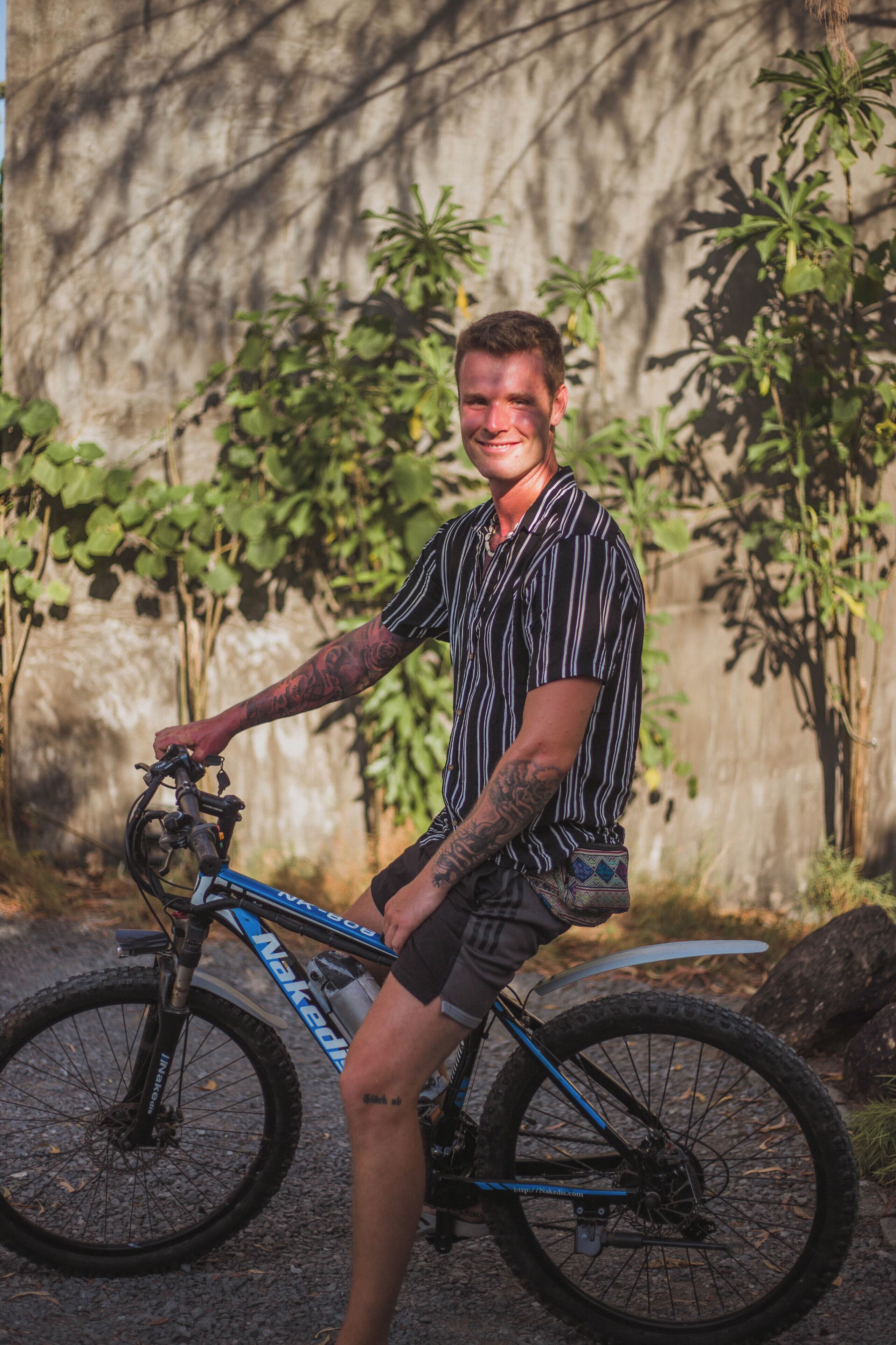 A person sitting on a blue mountain bike with a tattooed arm, wearing a striped shirt and shorts, in front of a stone wall with greenery.