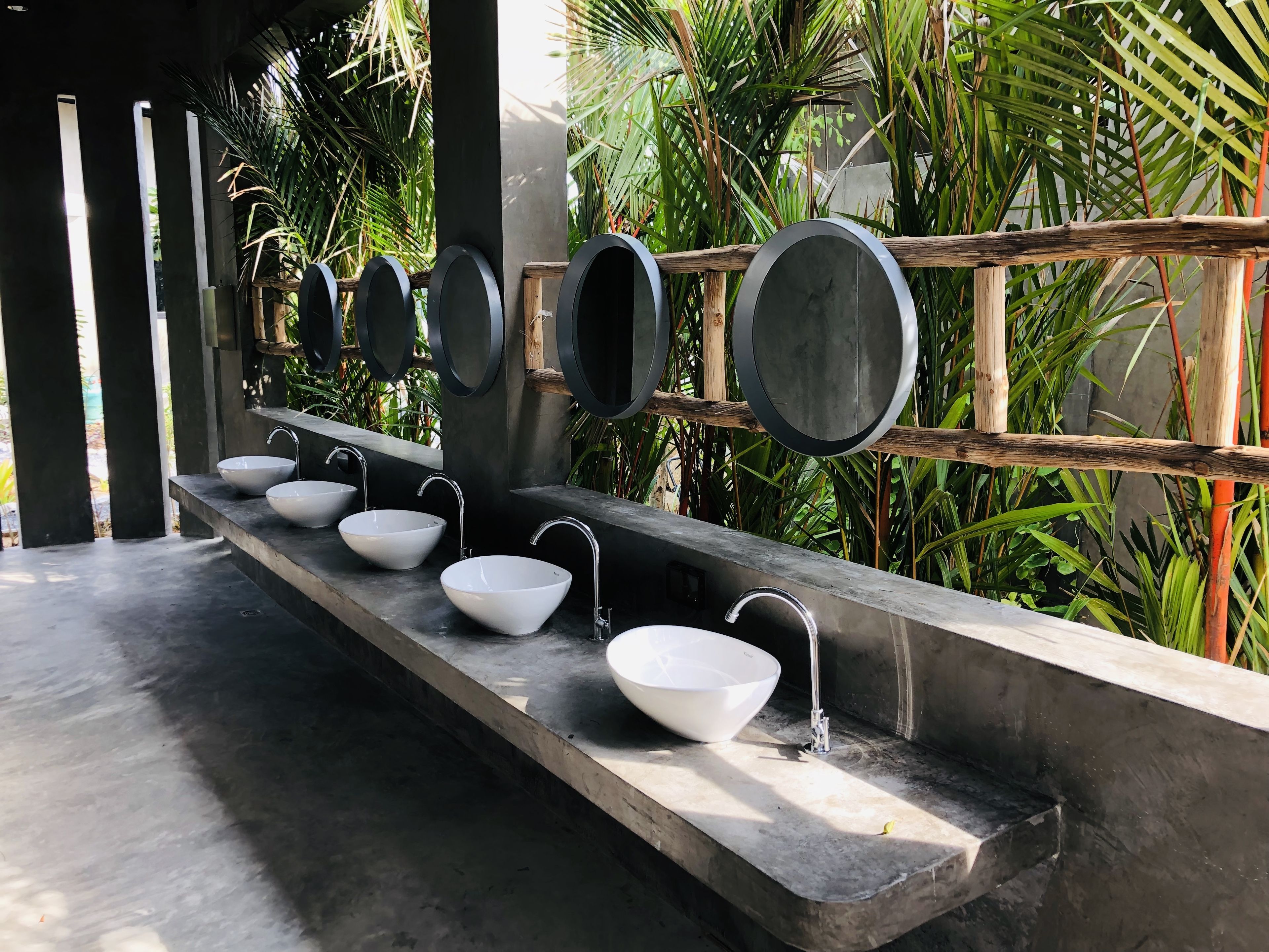 A row of white sinks with modern faucets, set against a backdrop of lush greenery and a concrete structure.