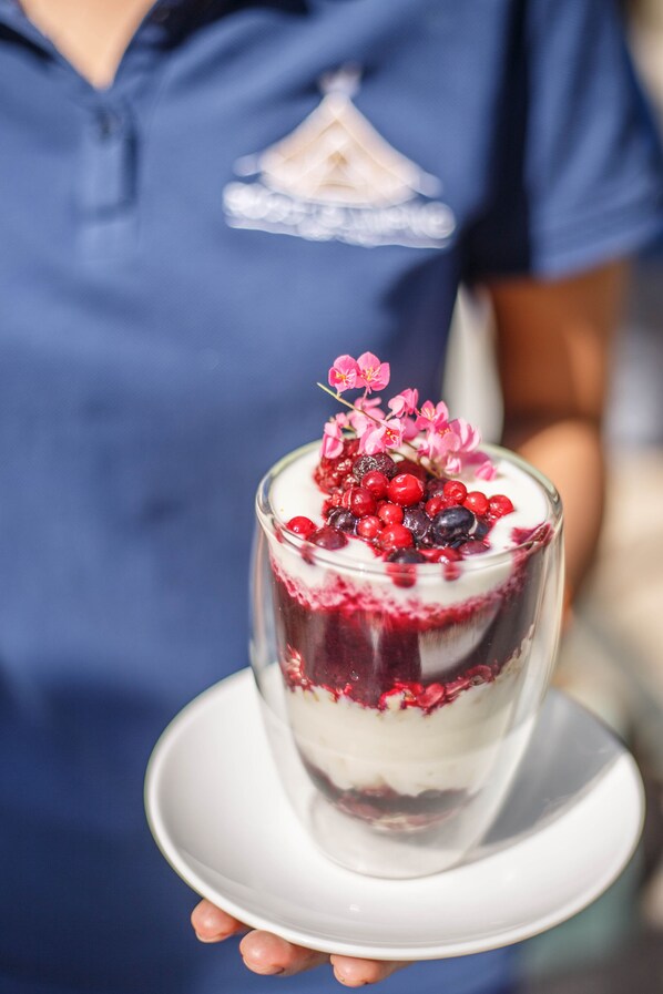 Sobremesa em copo transparente com frutas vermelhas e flores por cima, segurada por uma pessoa de camisa azul.